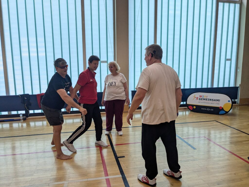 In einer Sporthalle befinden sich vier Personen vor einer Fensterwand. Eine Frau in roten T-Shirt wehrt den Angriff eines Mannes mit dunkler Brille ab, der sie von hinten umklammern will. Eine Frau und ein Mann in weißen T-Shirts stehen daneben bereit. Im Hintergrund als Bodenaufsteller ein Banner mit dem Aufdruck " Wir! Gemeinsam!" 